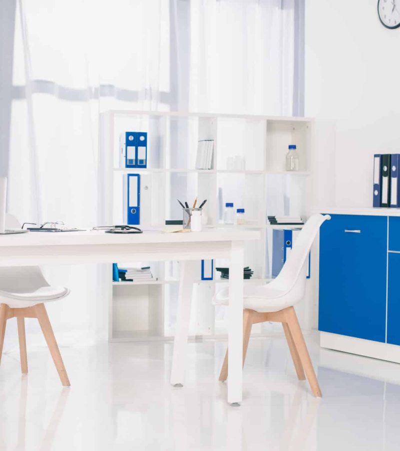 Bureau moderne avec du mobilier blanc et bleu, un bureau sur lequel se trouvent un ordinateur portable et des fournitures de bureau, des chaises, des étagères avec des classeurs et une horloge murale. Une lumière du soleil éclatante inonde la pièce.